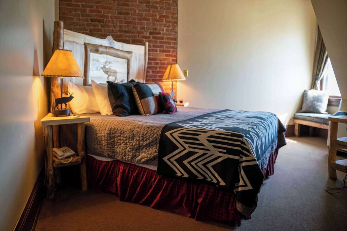 A bedroom with a brick wall, a bed with a patterned bedspread, two bedside lamps, a nightstand with a phone, and a chair by the window.
