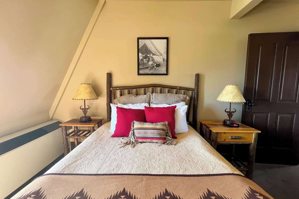 A neatly made bed with a patterned bedspread, two bedside lamps, a framed picture on the wall, and a wooden door.