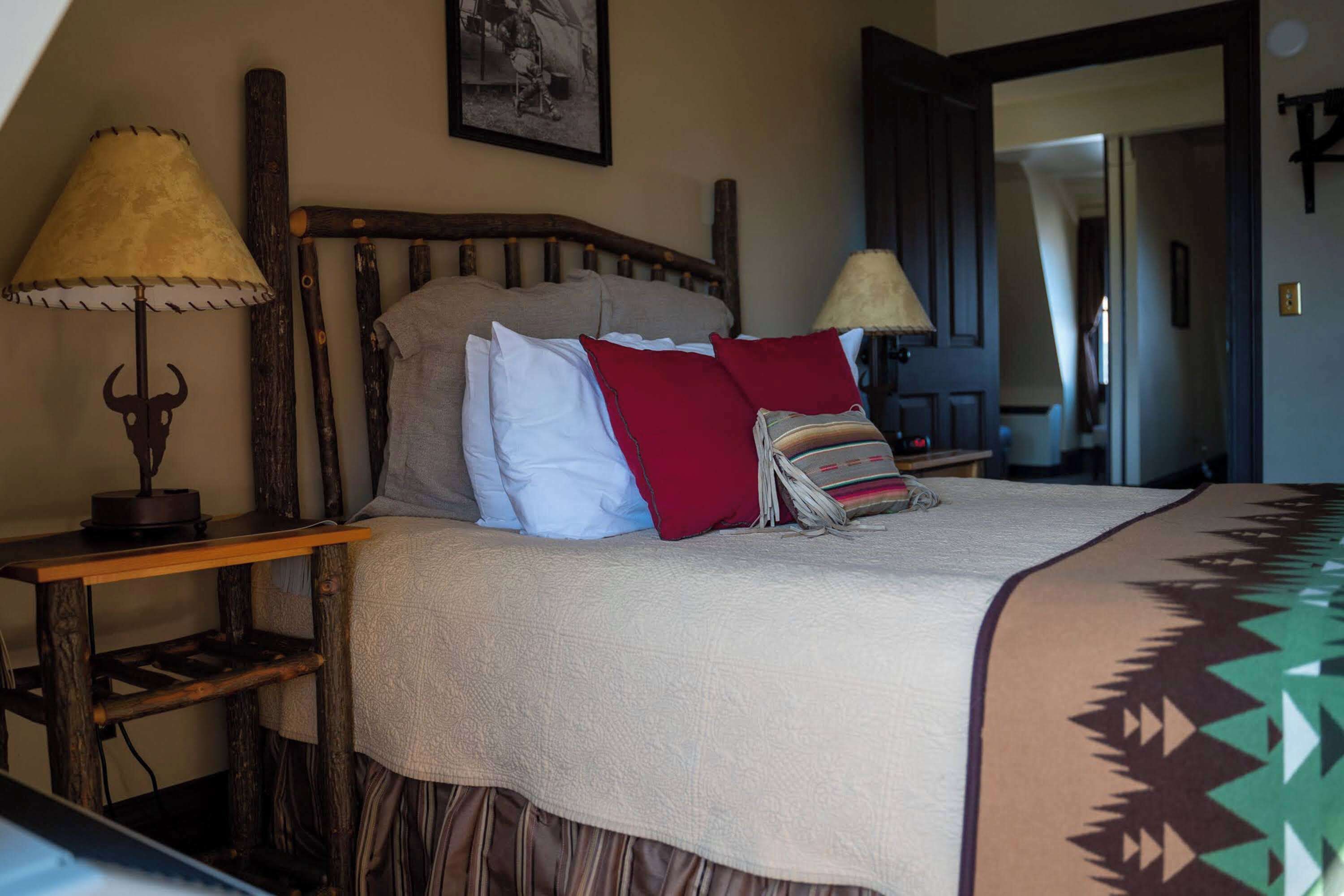 A bed with a wooden headboard, a beige blanket, and a patterned throw. There are two bedside tables with lamps.