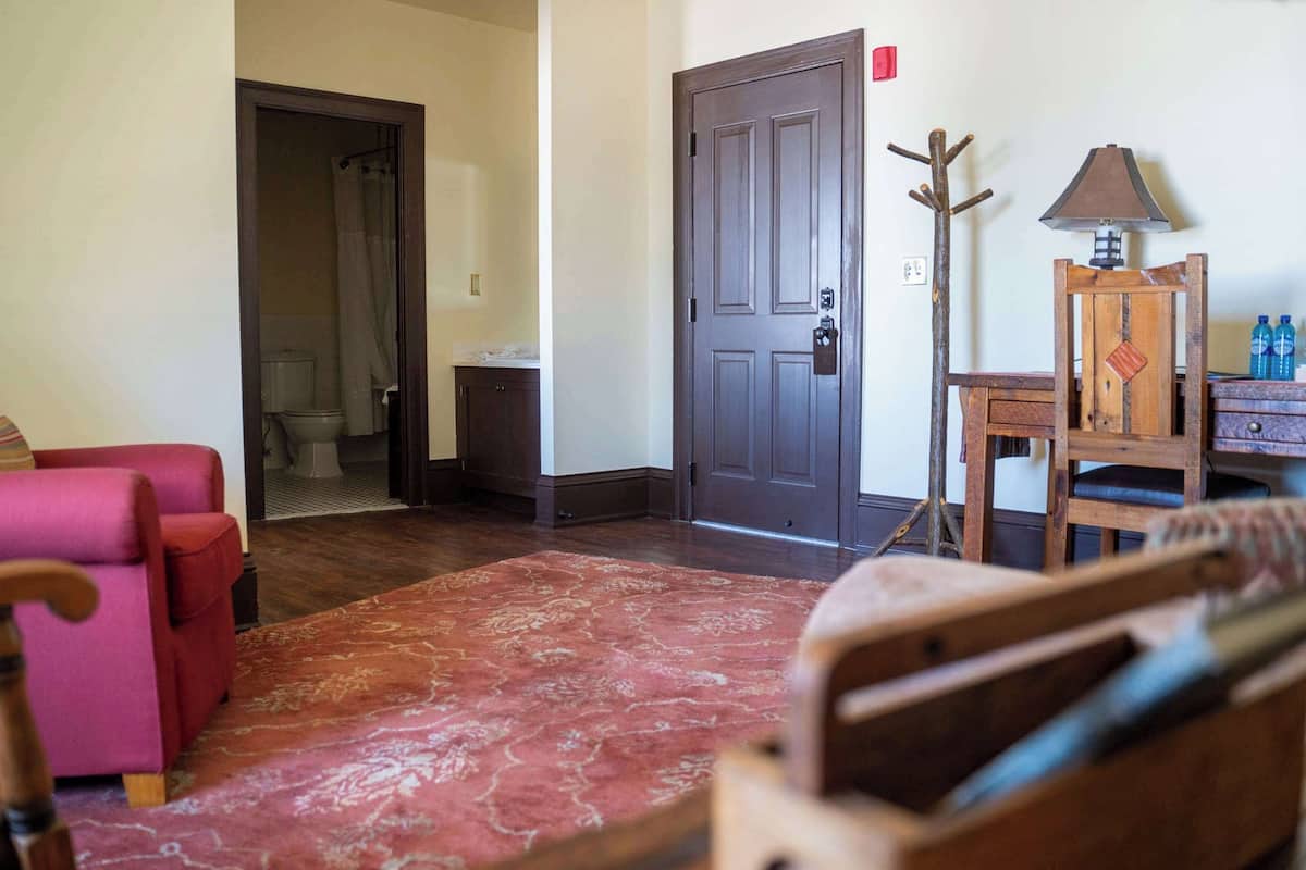 A room with a wooden door, a red patterned rug, a pink chair, and a wooden cabinet with a lamp.