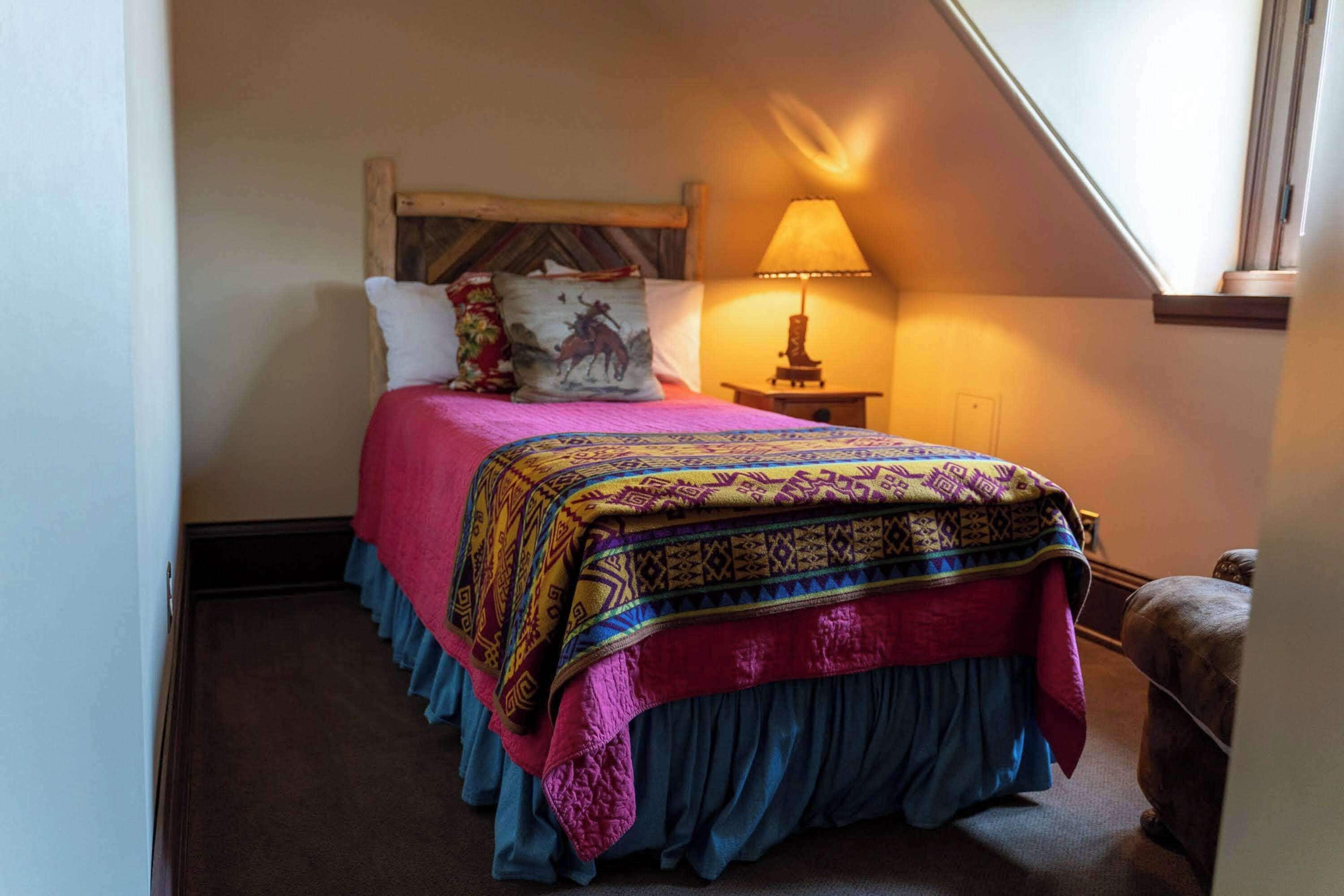 A bedroom with a bed covered in a colorful blanket, a wooden headboard, a bedside lamp, and a window with a view of the outside.
