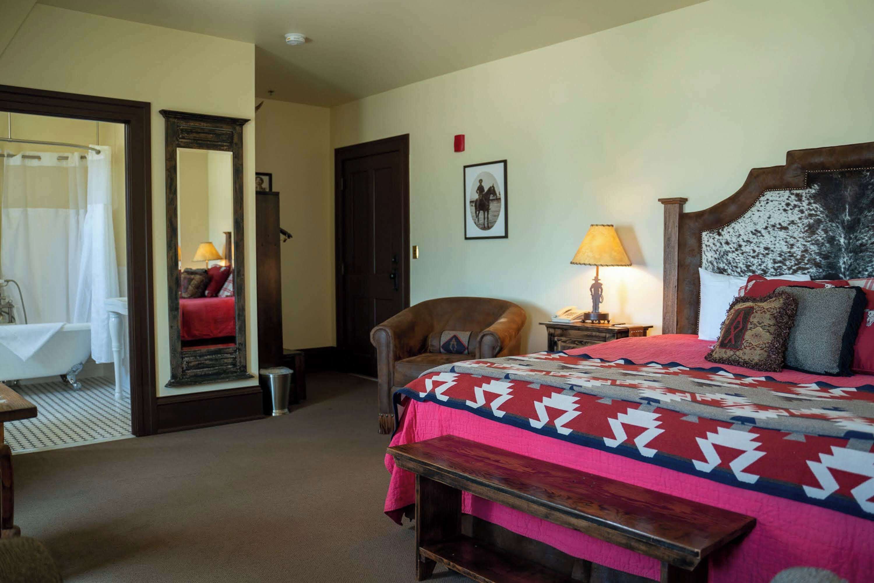 Une chambre d’hôtel comprenant un lit, une chaise, une table de chevet avec une lampe, un miroir et une salle de bain avec une baignoire et un rideau de douche.