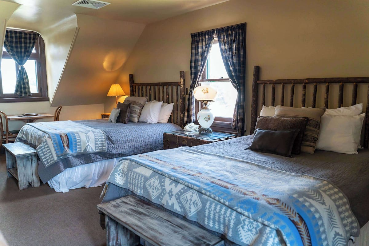 A room with two beds, a wooden headboard, a bedside table with a lamp, and a window with curtains.