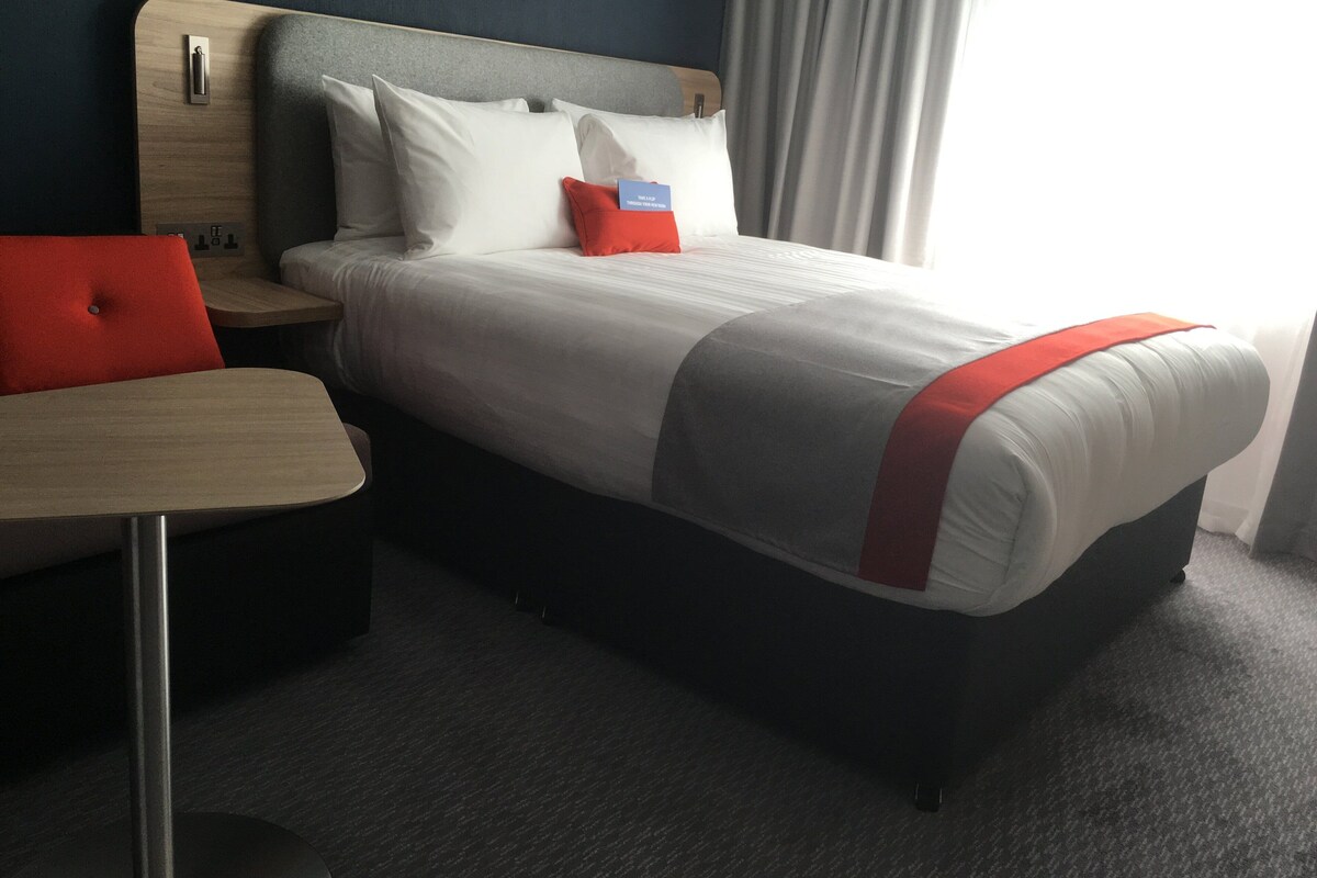 Une chambre d’hôtel avec un lit, une table de chevet, un bureau et une fenêtre avec des rideaux.