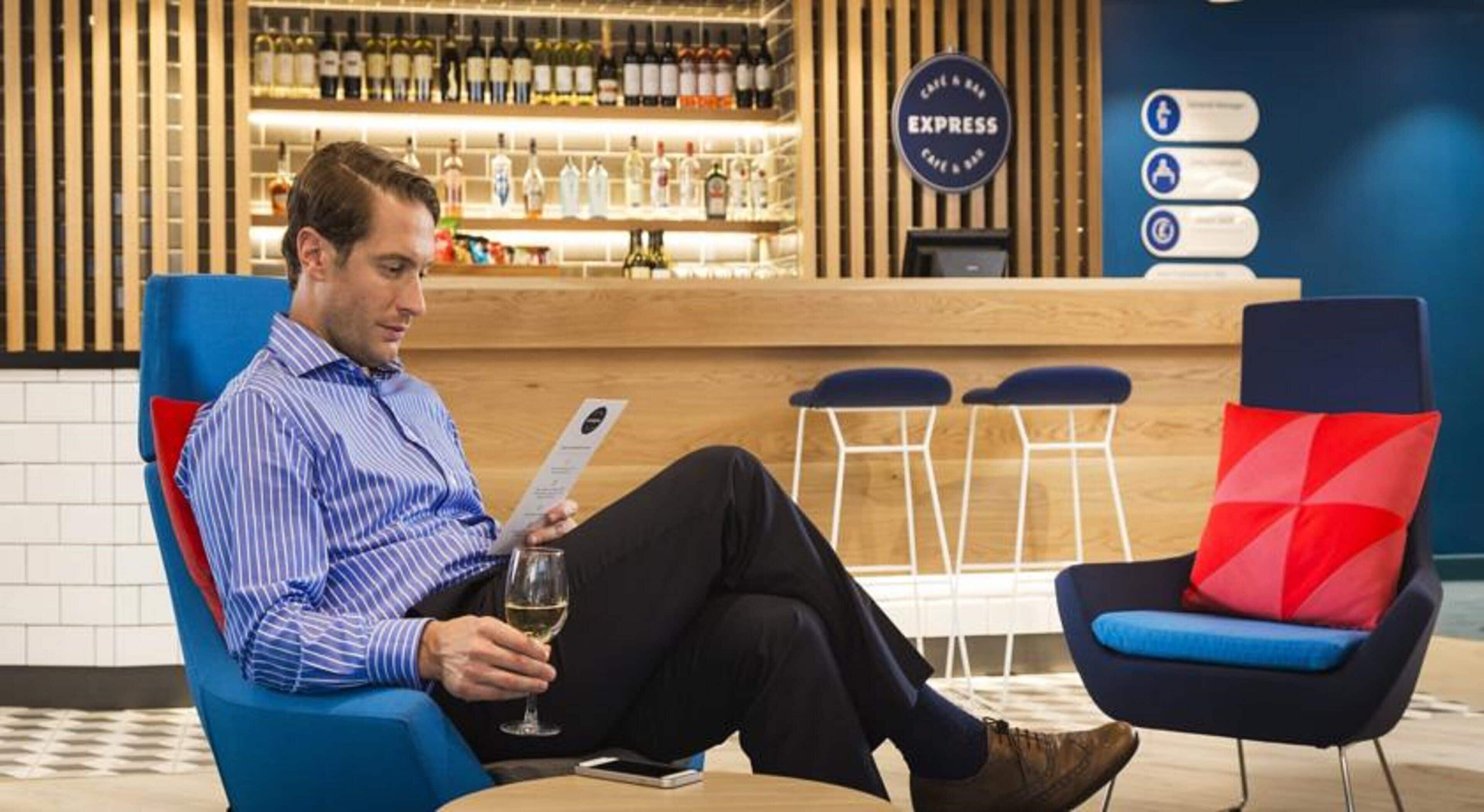 A man seated in a blue armchair with a red cushion, holding a glass of wine and a smartphone. In the background, there is a bar area with a sign that reads 'EXPRESS' and a menu board.