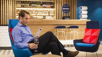 A man seated in a blue armchair with a red cushion, holding a glass of wine and a smartphone. In the background, there is a bar area with a sign that reads 'EXPRESS' and a menu board.