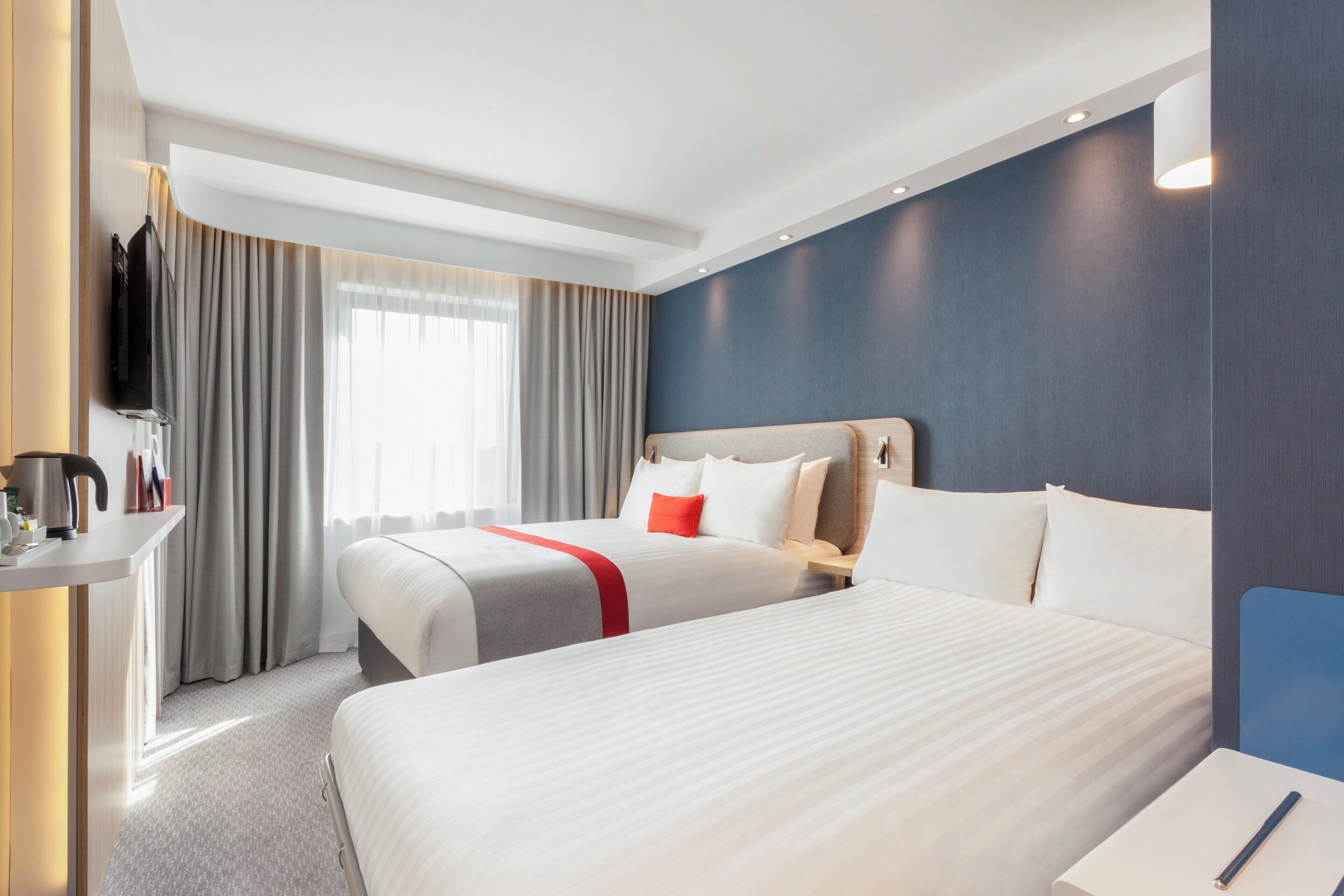 A hotel room with two beds, a TV, a desk, and a window with curtains.
