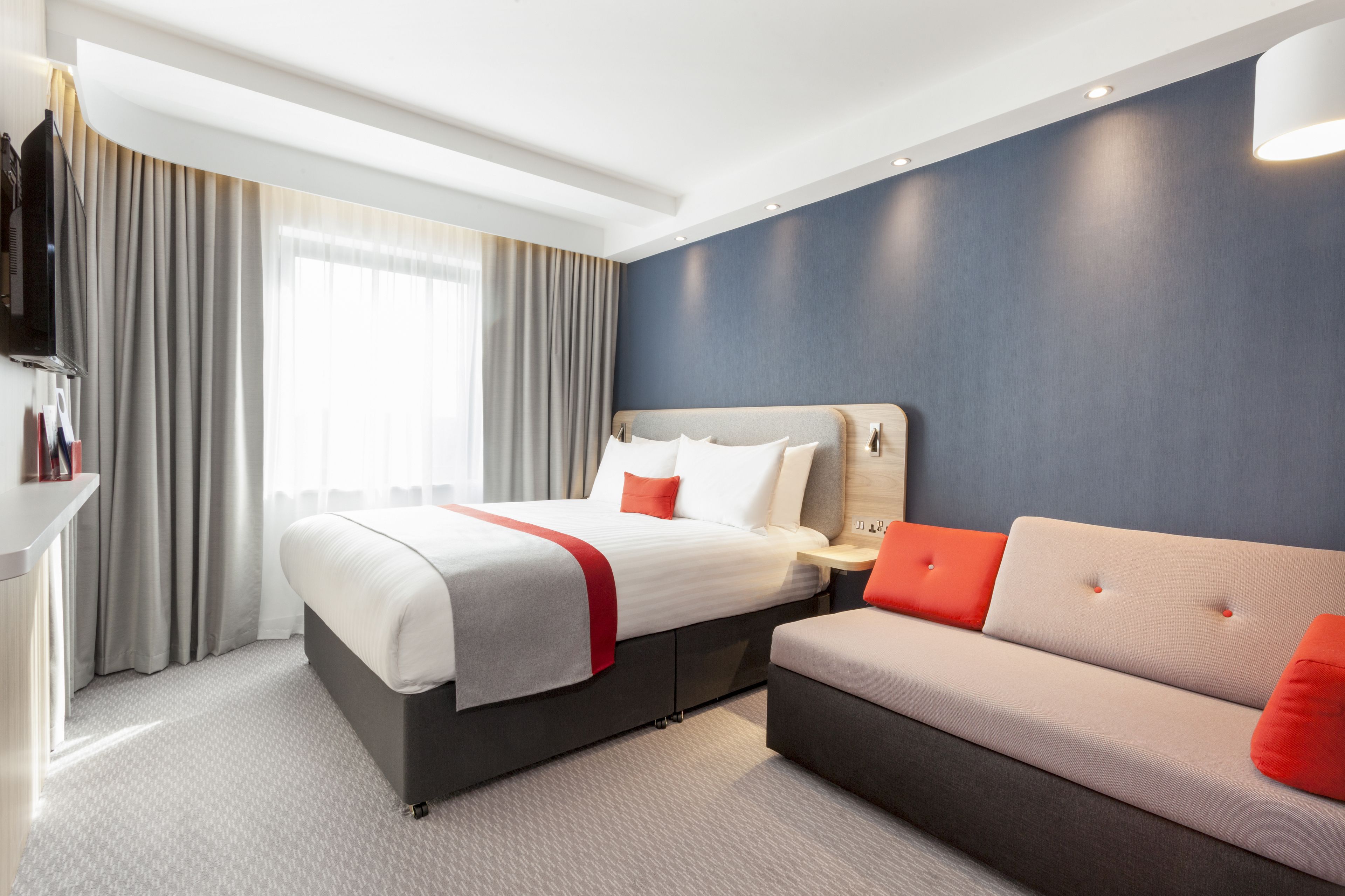 A modern hotel room with a large bed, a sofa, a flat-screen TV, and a window with curtains.