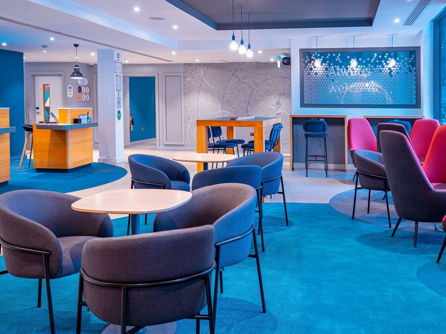 A modern hotel lobby with blue carpet, multiple seating areas, a reception desk, and a large window with a geometric design.
