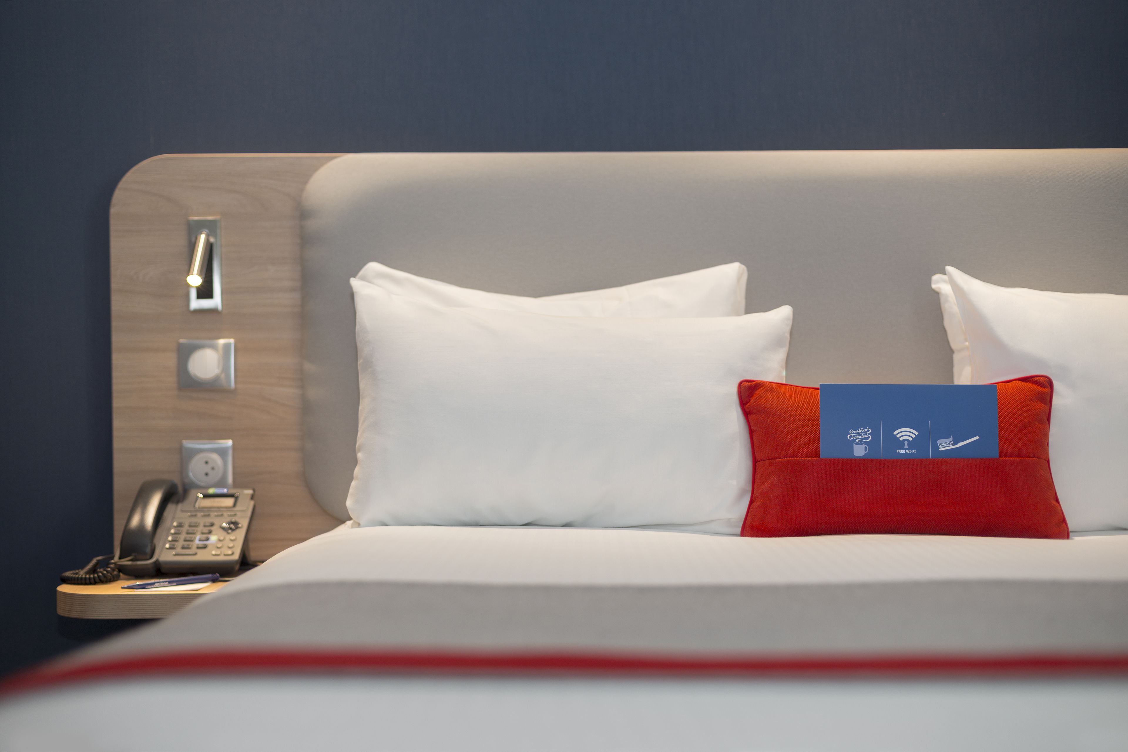 Une chambre d’hôtel avec un lit, des oreillers blancs, un oreiller rouge avec une étiquette bleue, un téléphone sur la table de chevet et une tête de lit en bois.