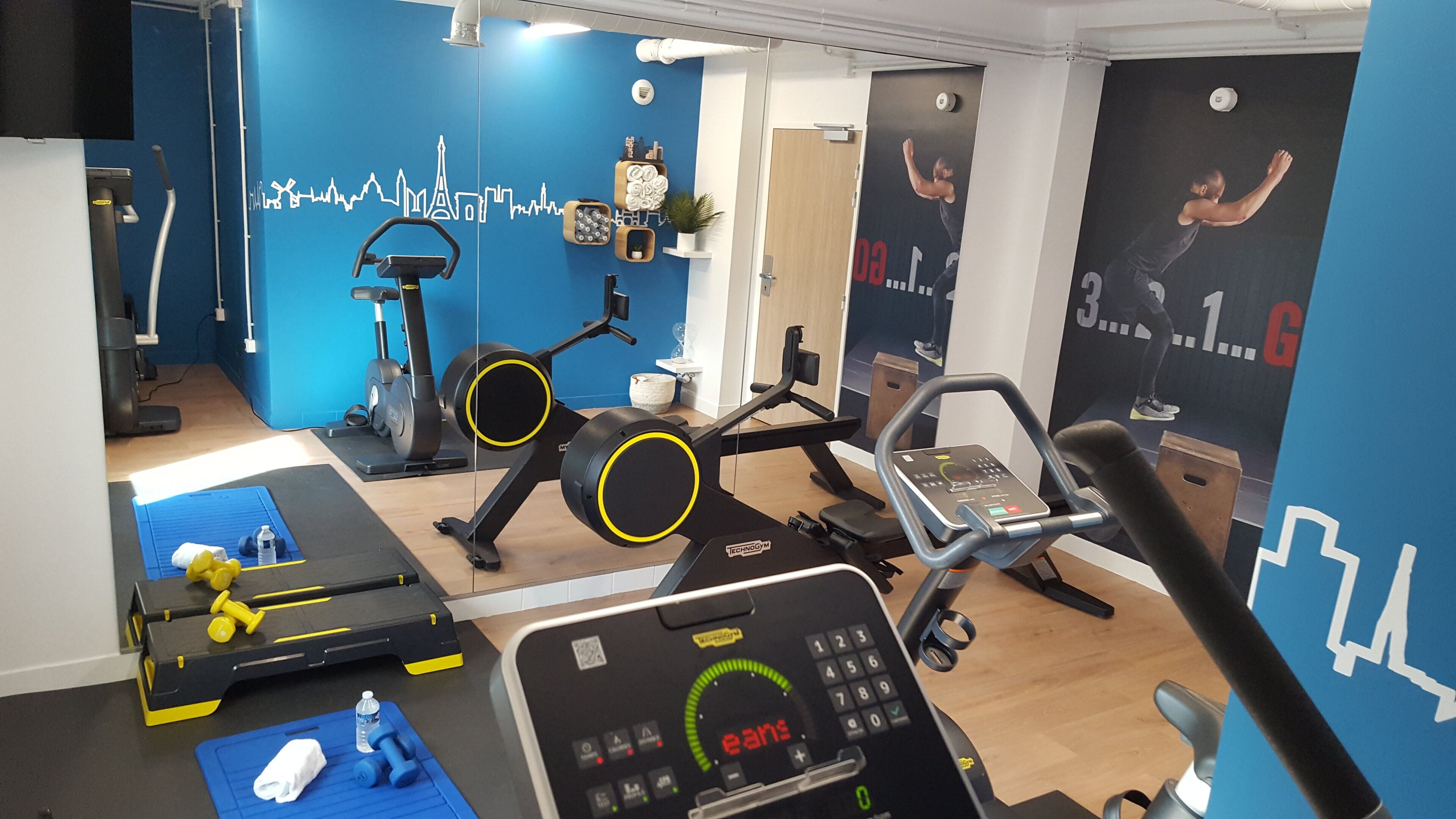 A gym with exercise equipment, a blue wall with a city skyline mural, and a wooden floor.