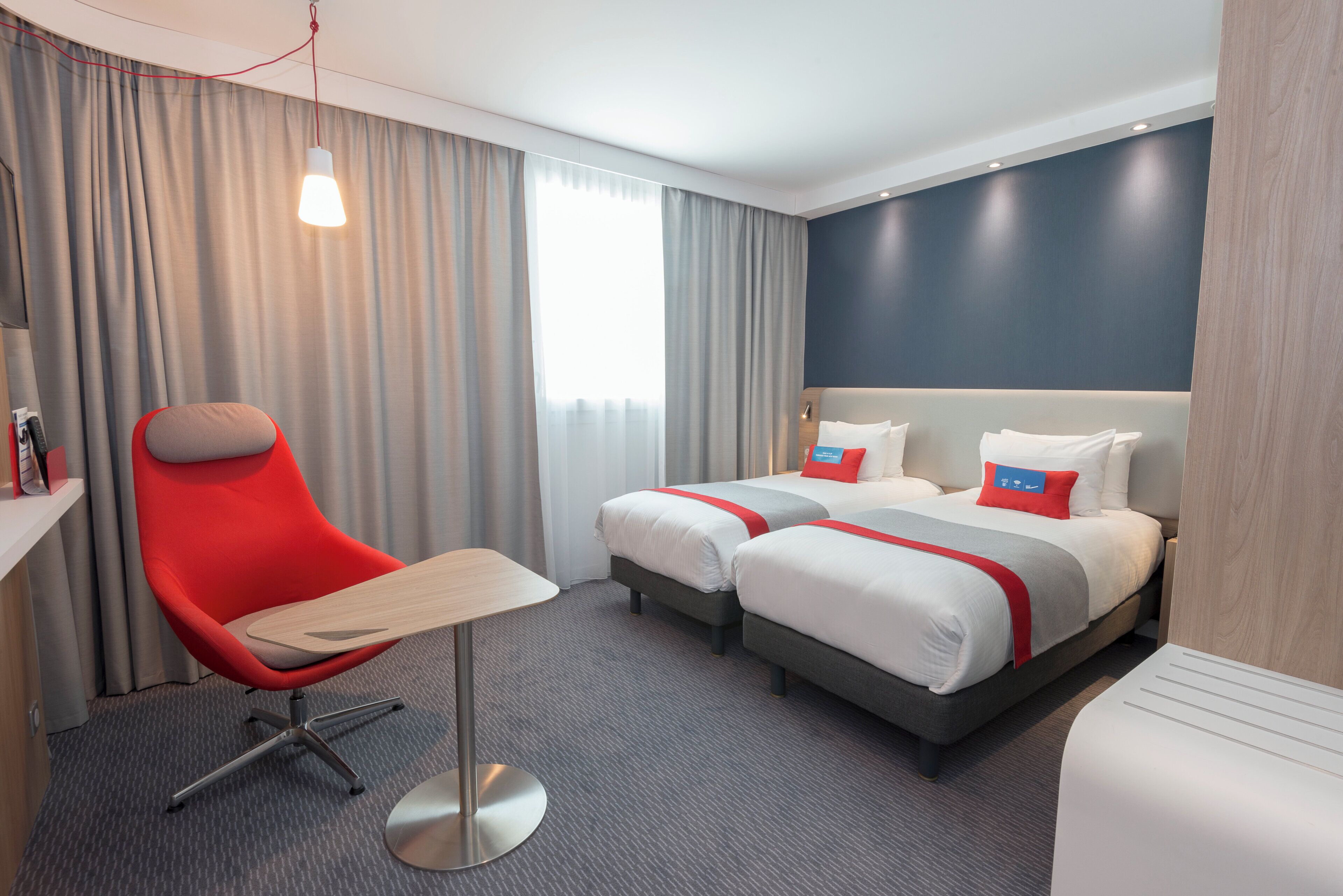 Een hotelkamer met twee bedden, een bureau, een stoel en een groot raam met gordijnen.