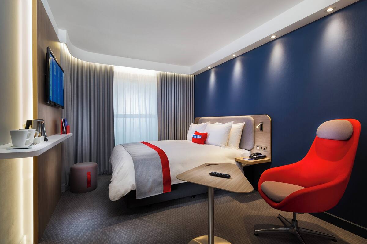 A modern hotel room with a bed, a red chair, a desk with a remote control, and a TV on the wall.