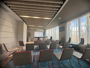 Un salón de conferencias con diseño moderno, que cuenta con una pantalla grande, varias sillas y vistas al exterior a través de amplios ventanales.