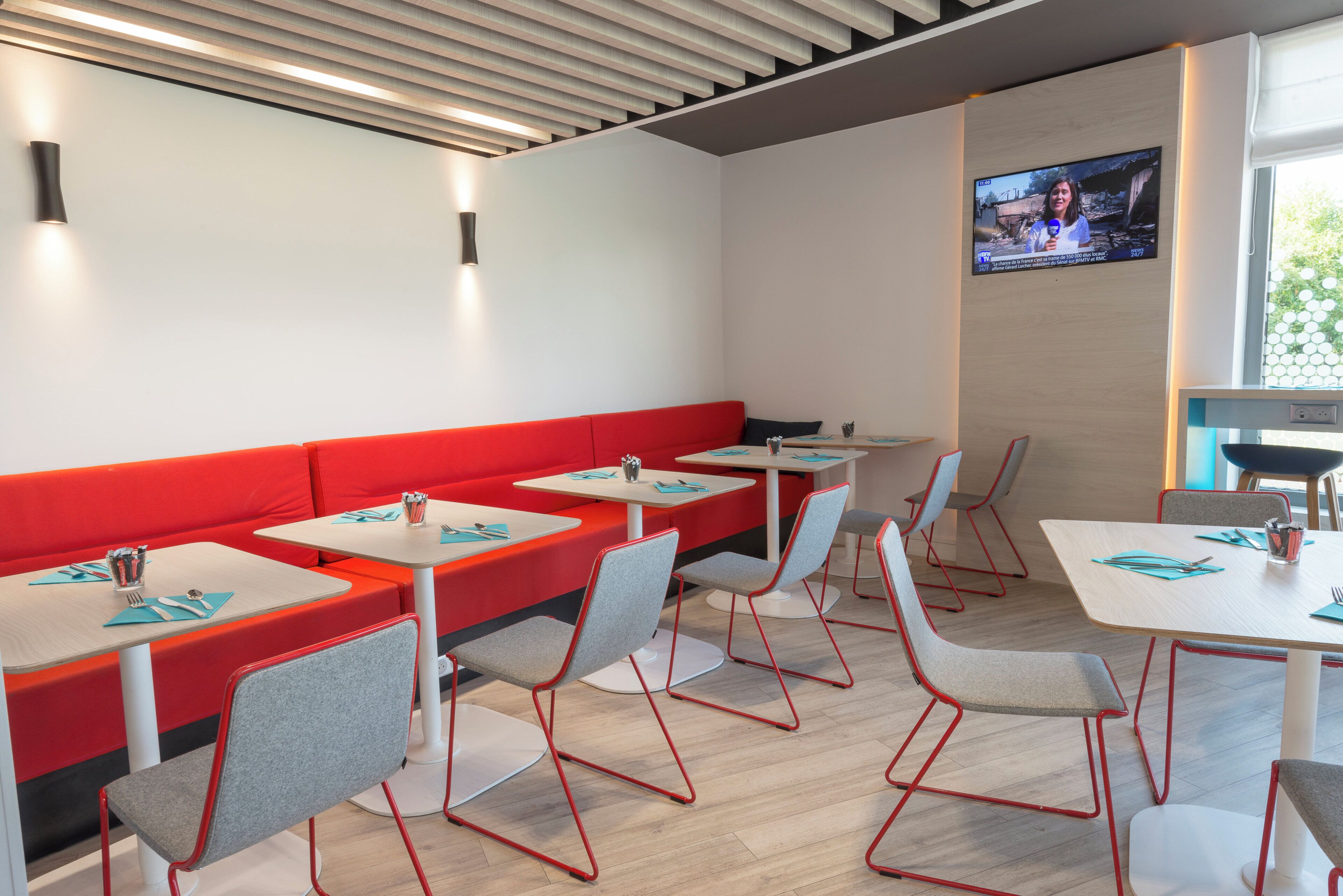 A modern dining area with red booths and grey chairs, a flat-screen TV mounted on the wall, and a window providing natural light.