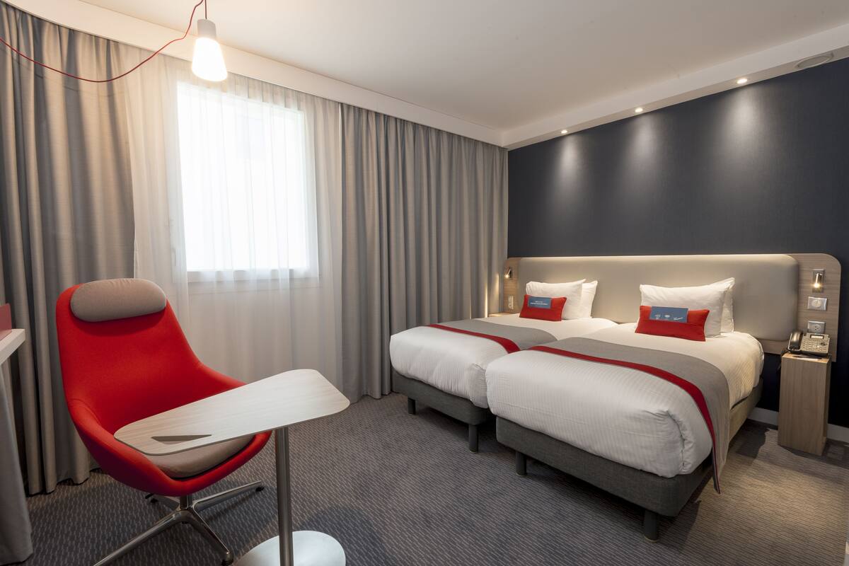 A hotel room with a bed, two bedside tables, a red chair, a desk, and a window with curtains.