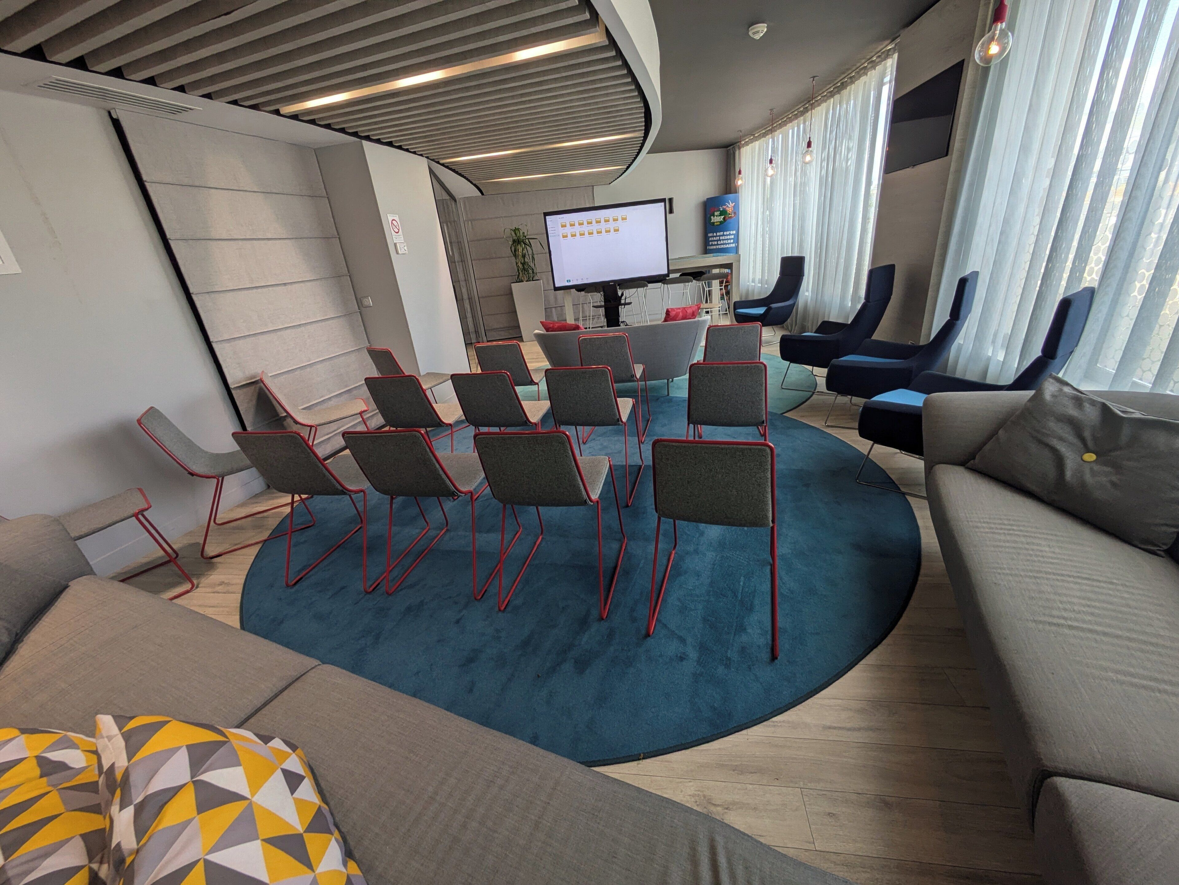 Une salle de conférence moderne équipée d’un grand écran, de nombreuses chaises, d’un canapé et d’une moquette bleue.