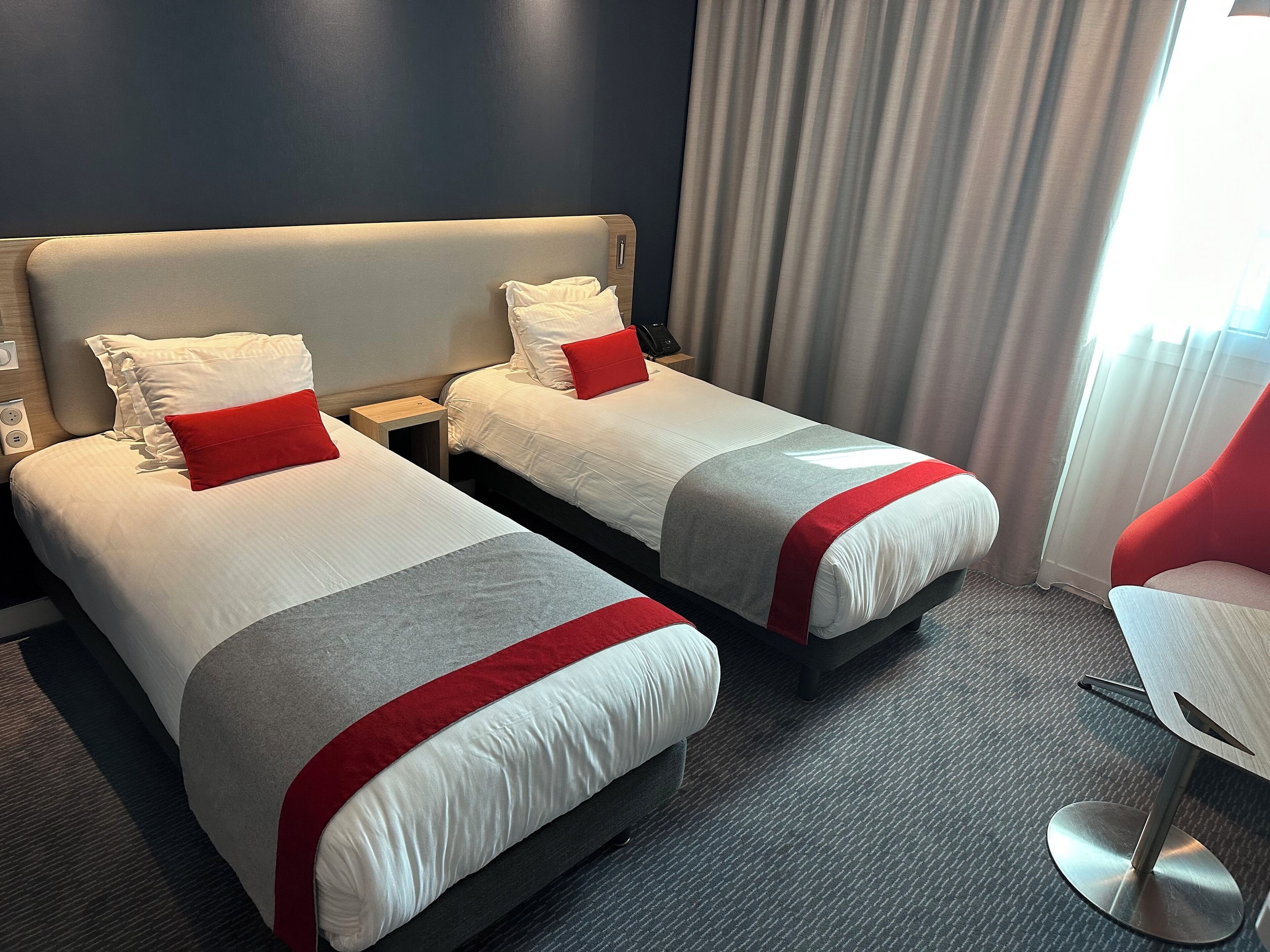 A hotel room with two beds, each with white bedding and red accents, a small bedside table, and a chair.