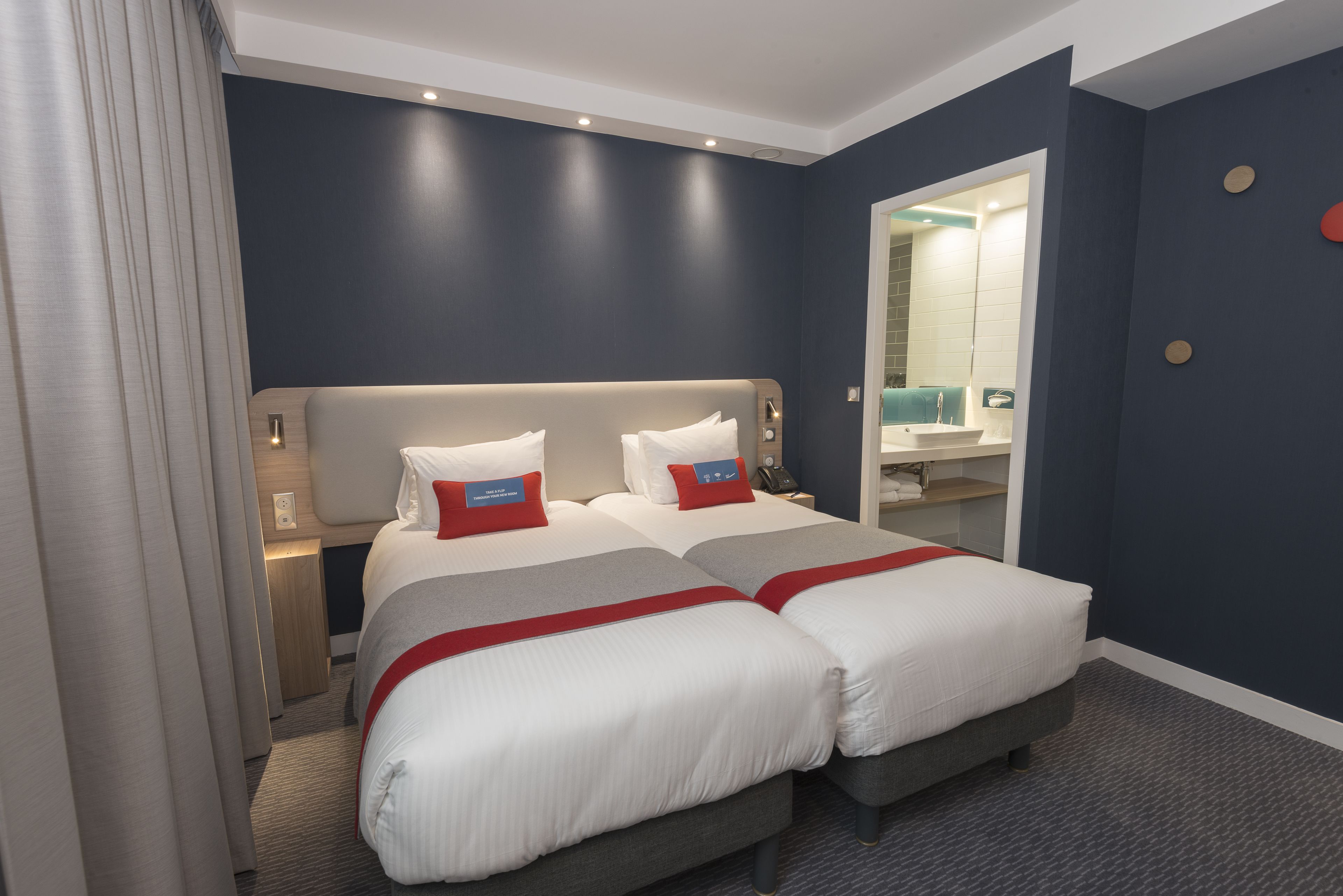 Une chambre d’hôtel avec deux lits, une salle de bain avec un lavabo et un miroir, et une porte donnant sur une autre pièce.