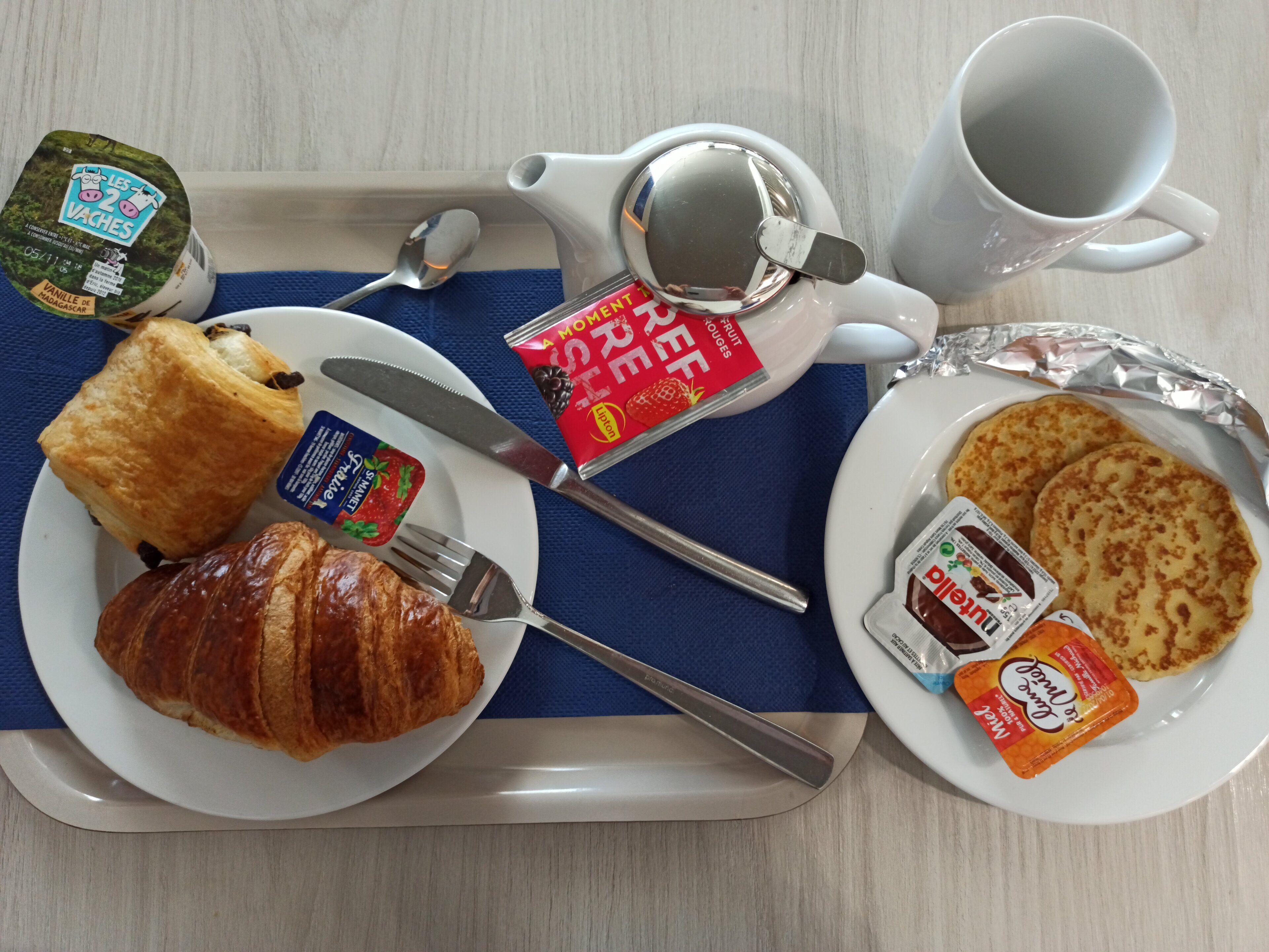 A breakfast tray with croissants, a jam packet, a mug, and a plate with pancakes and jam.