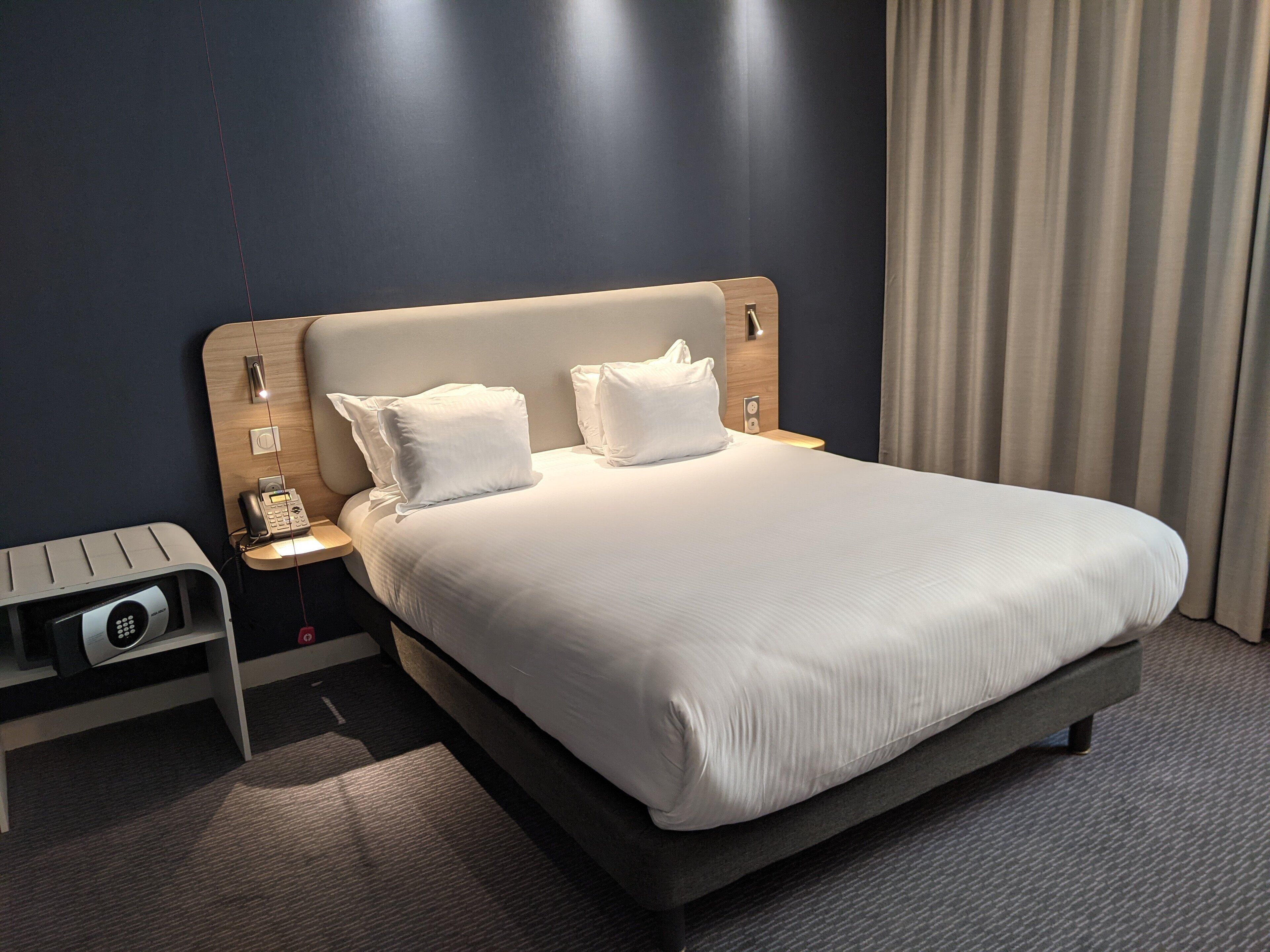 A modern hotel room with a large bed, two pillows, a bedside table with a phone, and a small desk with a computer.