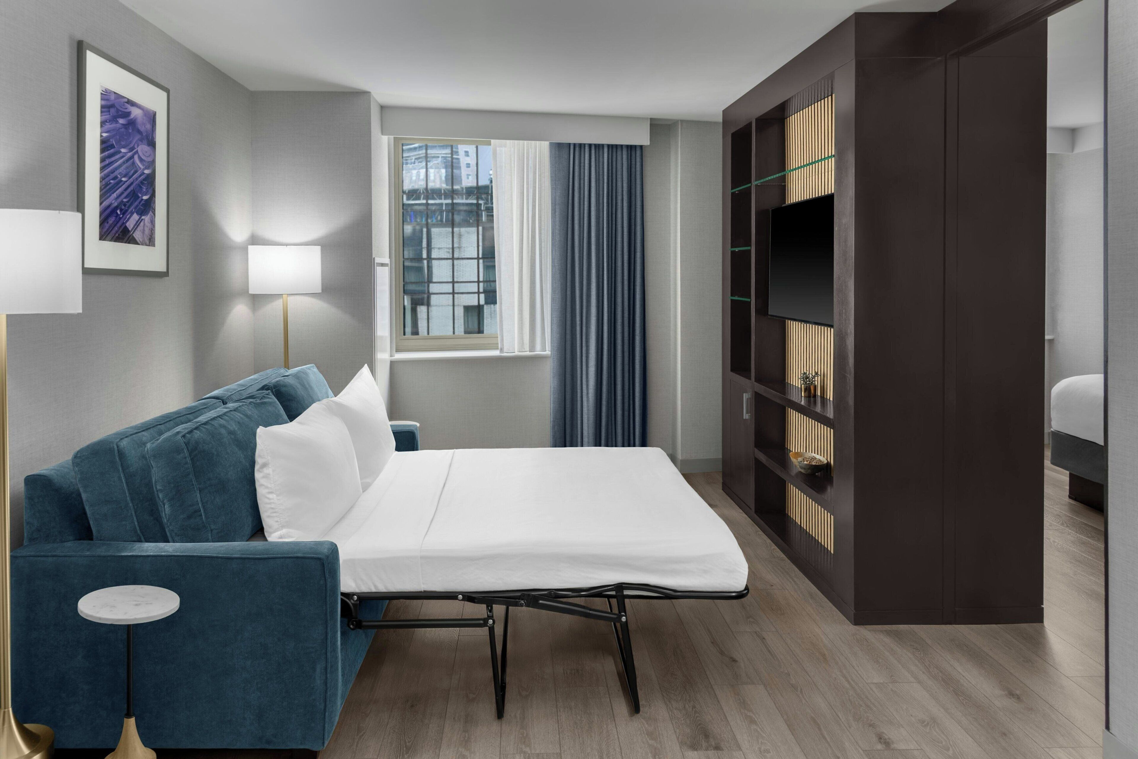 Een moderne hotelkamer met een blauwe bank, een bed met een wit matras, een klein rond tafeltje, een vloerlamp, een raam met gordijnen en een ingebouwde tv-kast.