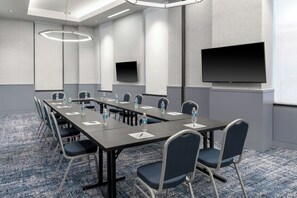 Une salle de conférence équipée d’une longue table, de chaises, de bouteilles d’eau, d’un écran de projection et d’un téléviseur à écran plat.