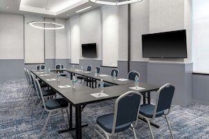 Uma sala de conferência com uma mesa comprida, cadeiras, garrafas de água, uma tela de projetor e uma TV de tela plana.