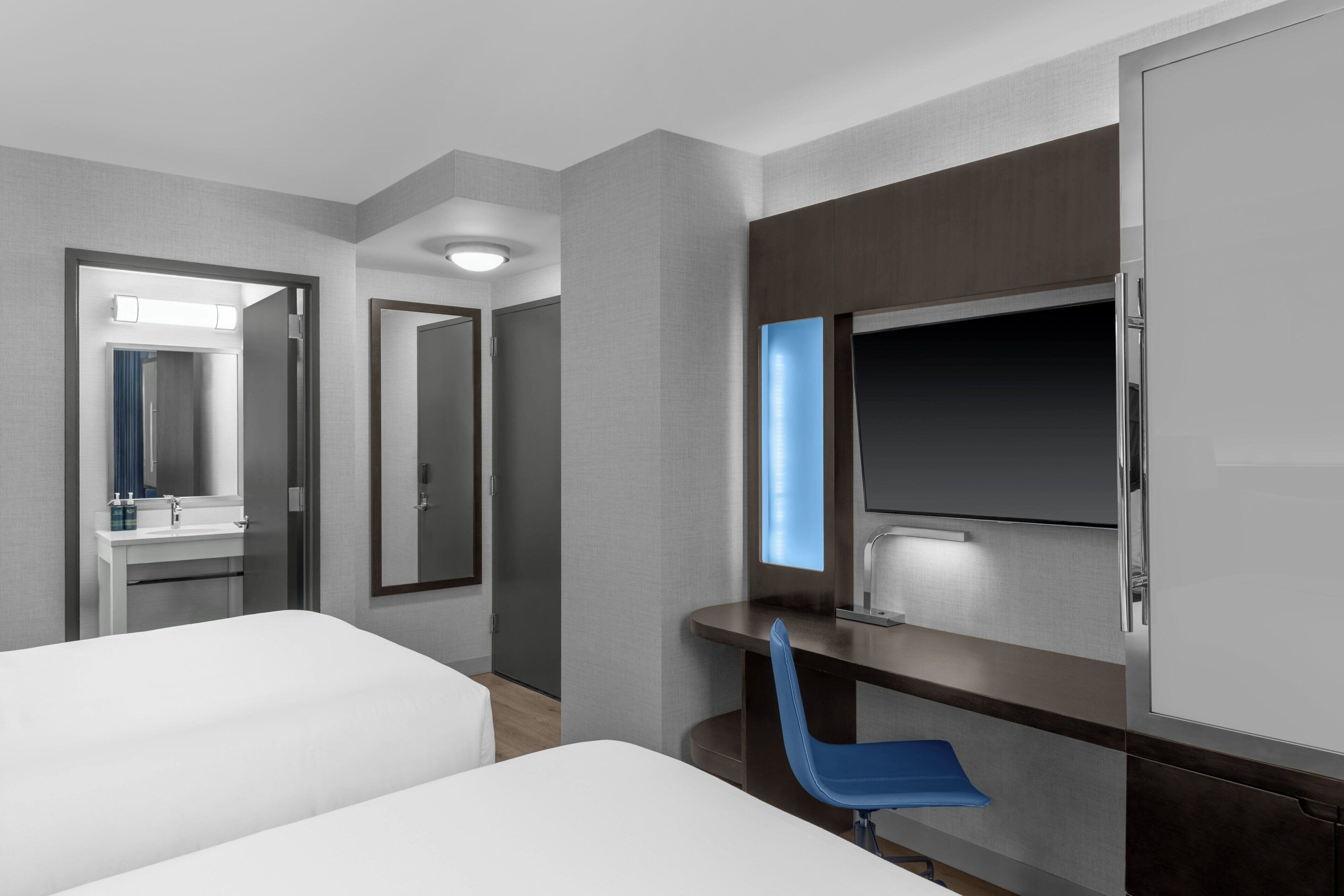 A hotel room with two beds, a desk, a mirror, and a large window.