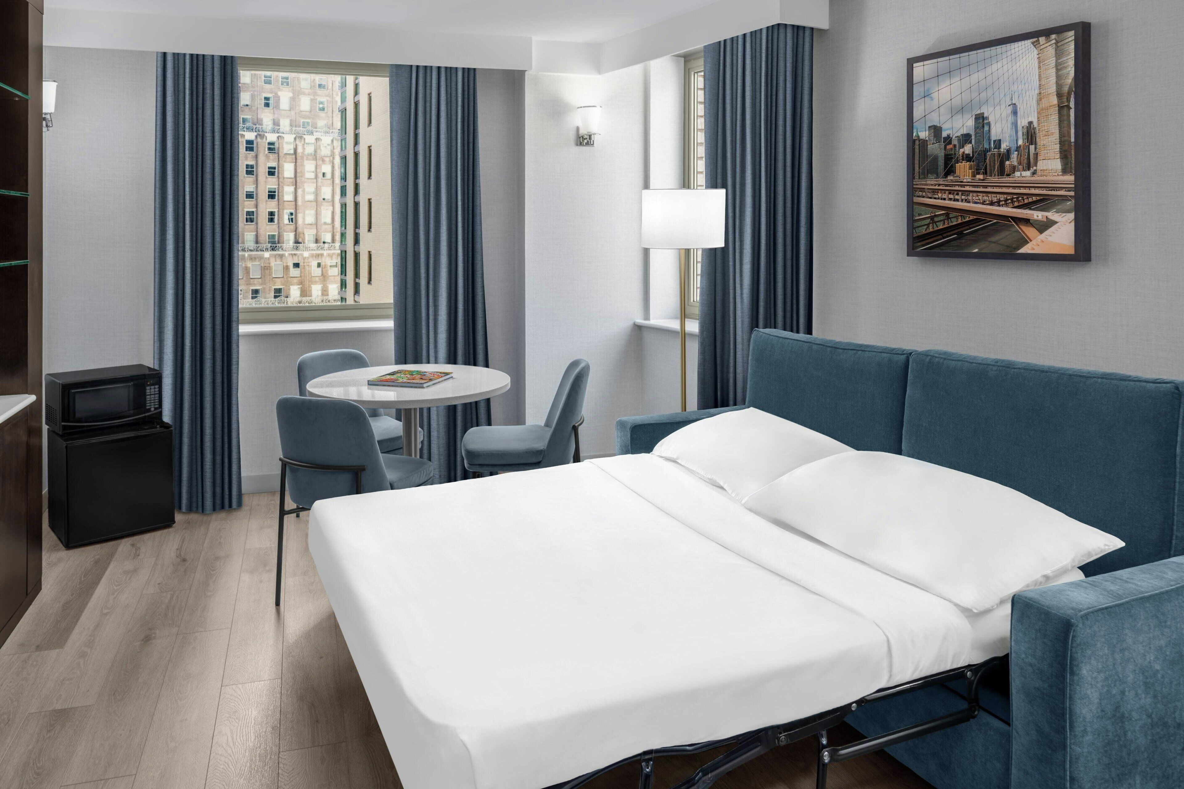 Habitación de hotel con cama, escritorio, silla, lámpara y una pintura de un paisaje urbano.
