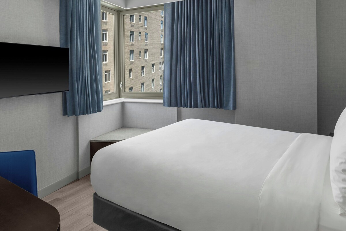 Une chambre d’hôtel comprenant un lit, un bureau, une chaise, un téléviseur fixé au mur et une fenêtre avec des rideaux.
