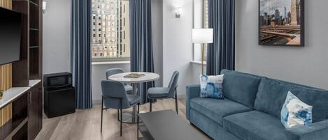 A modern hotel room with a sofa, dining table, chairs, a TV, a microwave, and a window with curtains.