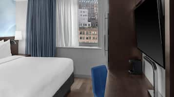 Een hotelkamer met een bed, een bureau met een televisie, een stoel, een raam met gordijnen en uitzicht op gebouwen.