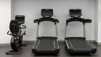 Three exercise machines, a stationary bike, and a treadmill in a room with a plain wall and a speckled floor.