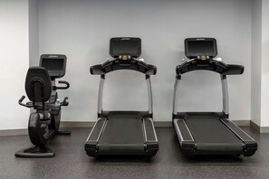 Dans une pièce aux murs unis et au sol à motifs, on trouve trois appareils d’exercice, dont un vélo d’appartement et un tapis de course.