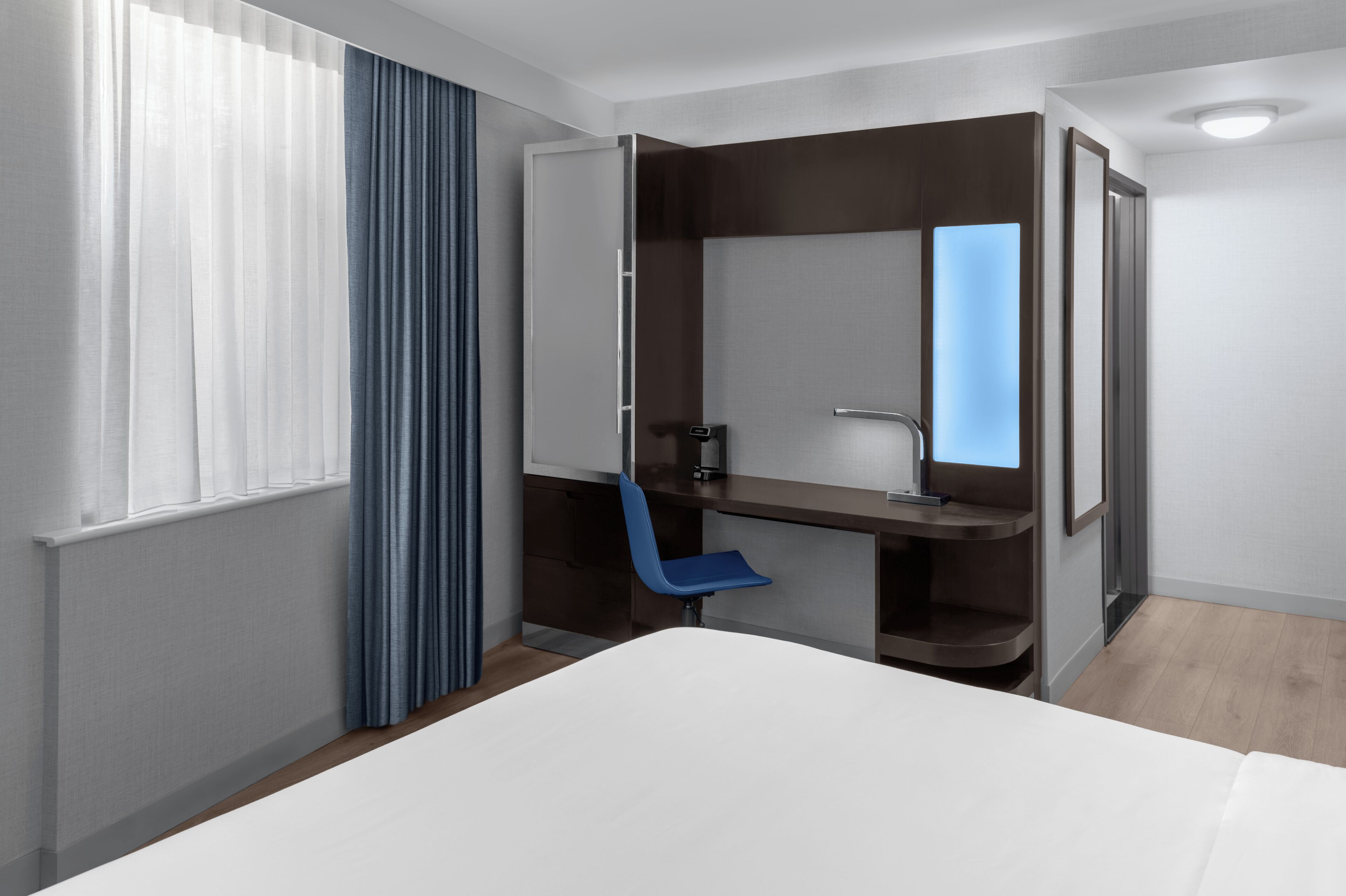 Une chambre d’hôtel moderne équipée d’un lit, d’un bureau, d’une chaise, d’un miroir et d’une fenêtre avec des rideaux.