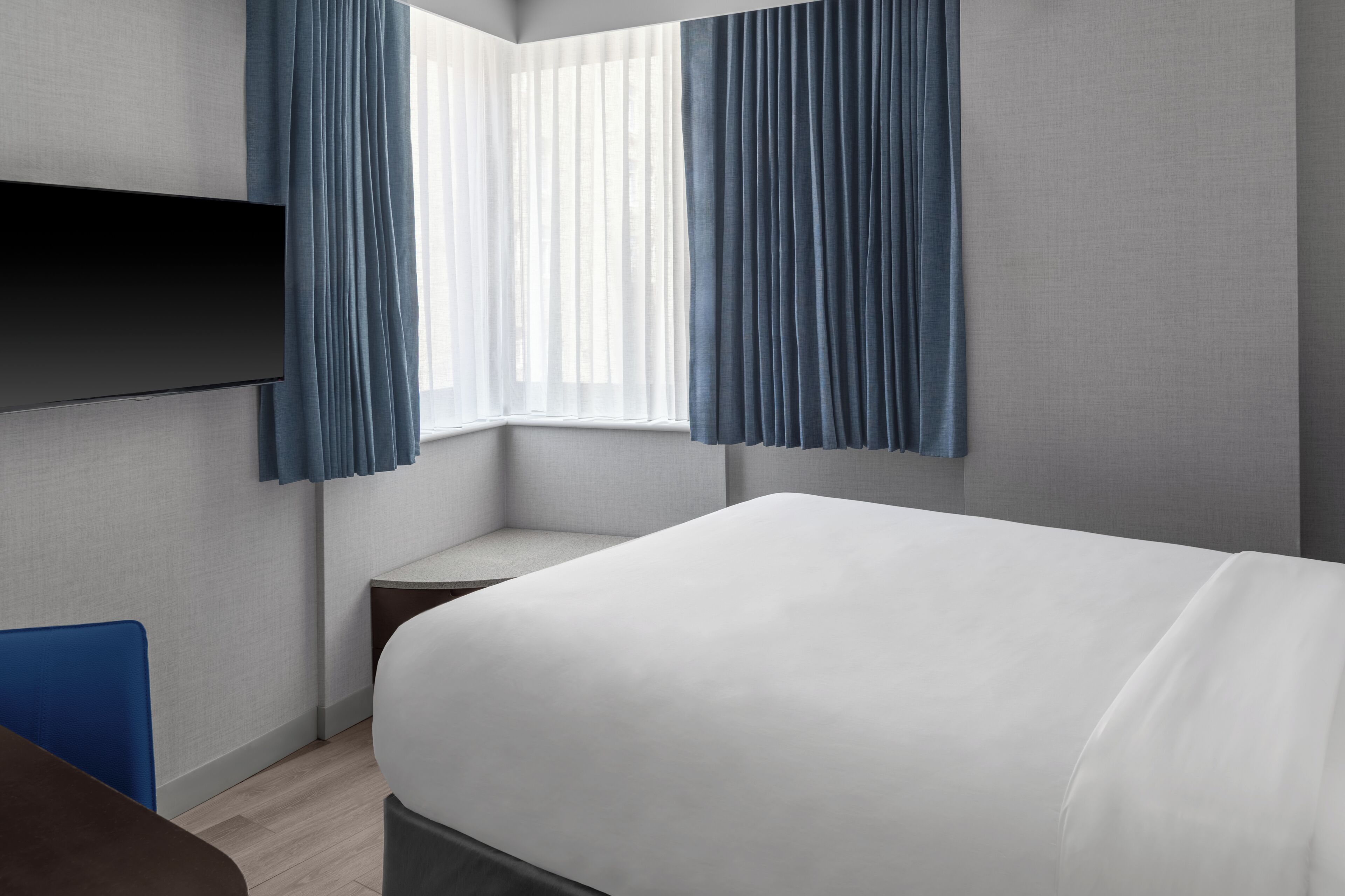 A modern hotel room with a large bed, a flat-screen TV mounted on the wall, and a window with blue curtains.