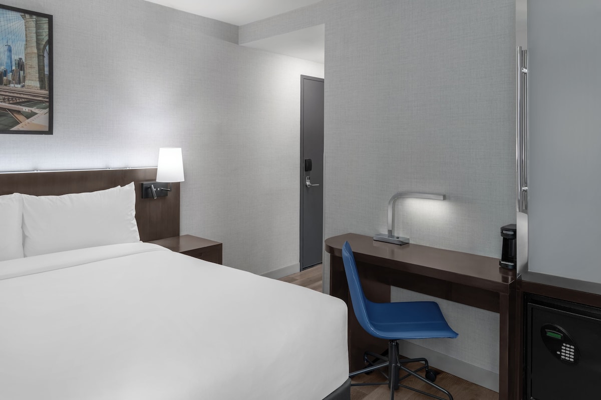 A hotel room with a bed, a desk with a chair, a nightstand with a lamp, a safe, and a door.