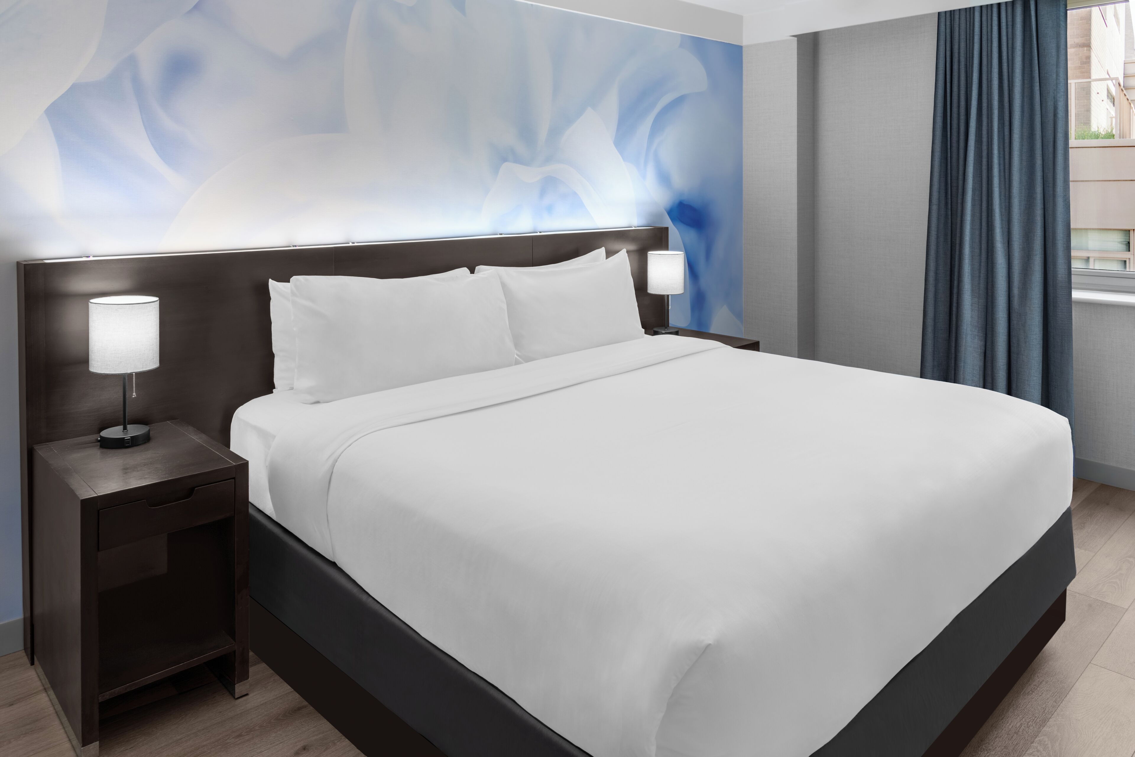 Una habitación de hotel moderna con una cama grande, dos mesitas de noche con lámparas, un cabecero oscuro y una pared con un diseño abstracto azul.