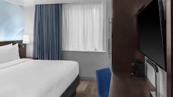 Ein Hotelzimmer mit Bett, Schreibtisch mit Flachbildfernseher, Stuhl, Fenster mit Vorhängen und einer Wandlampe.