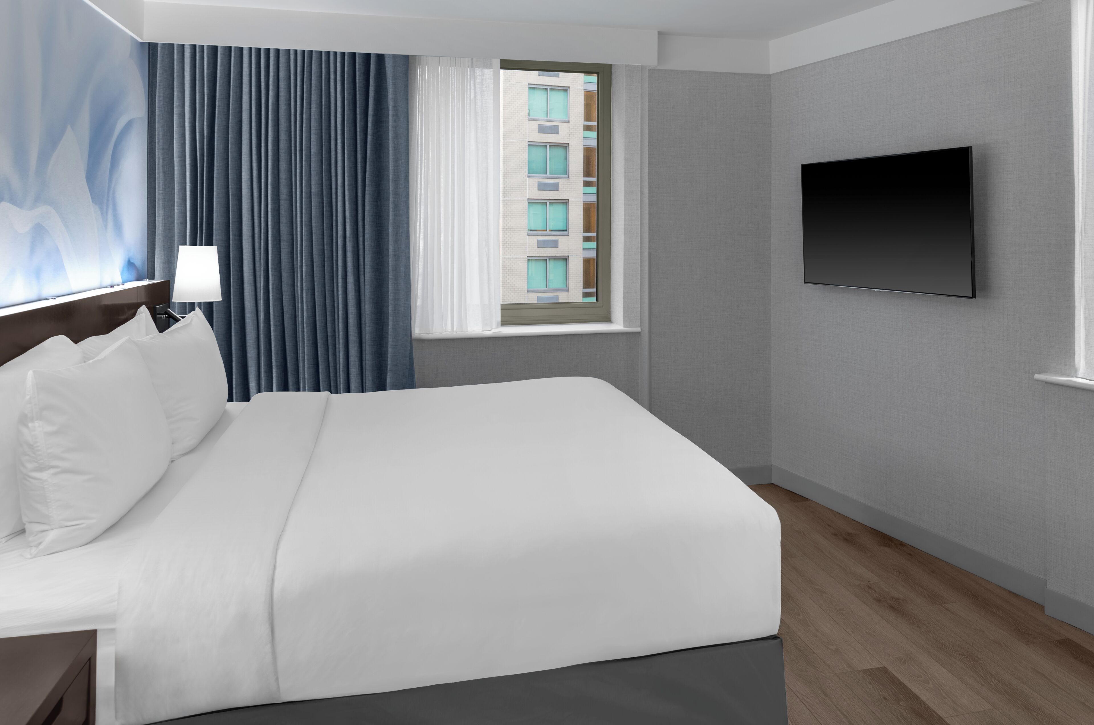 Habitación de hotel con cama, televisor de pantalla plana, ventana con cortinas, lámpara y vista a un edificio.