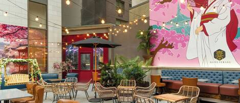 An outdoor seating area with wicker chairs and tables, a red umbrella, and a mural of a woman holding a fan.