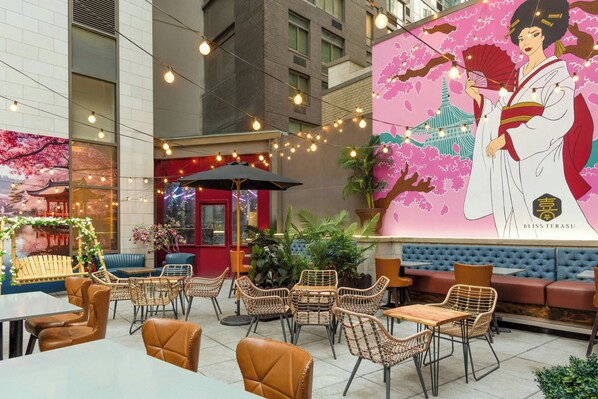 An outdoor seating area with wicker chairs and tables, a red umbrella, and a mural of a woman holding a fan. - Four Points By Sheraton New York Downtown (New York)