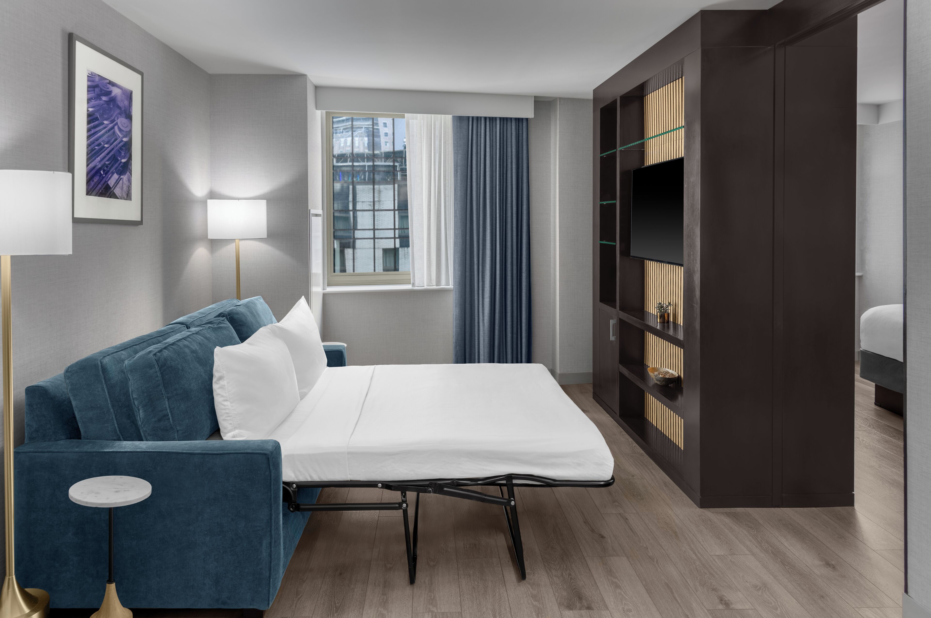 A hotel room with a bed, a blue sofa, a floor lamp, a window with curtains, and a built-in bookshelf.