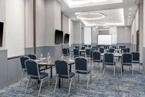 Un salón de conferencias con varias mesas y sillas, botellas de agua sobre las mesas y una pantalla grande al frente.