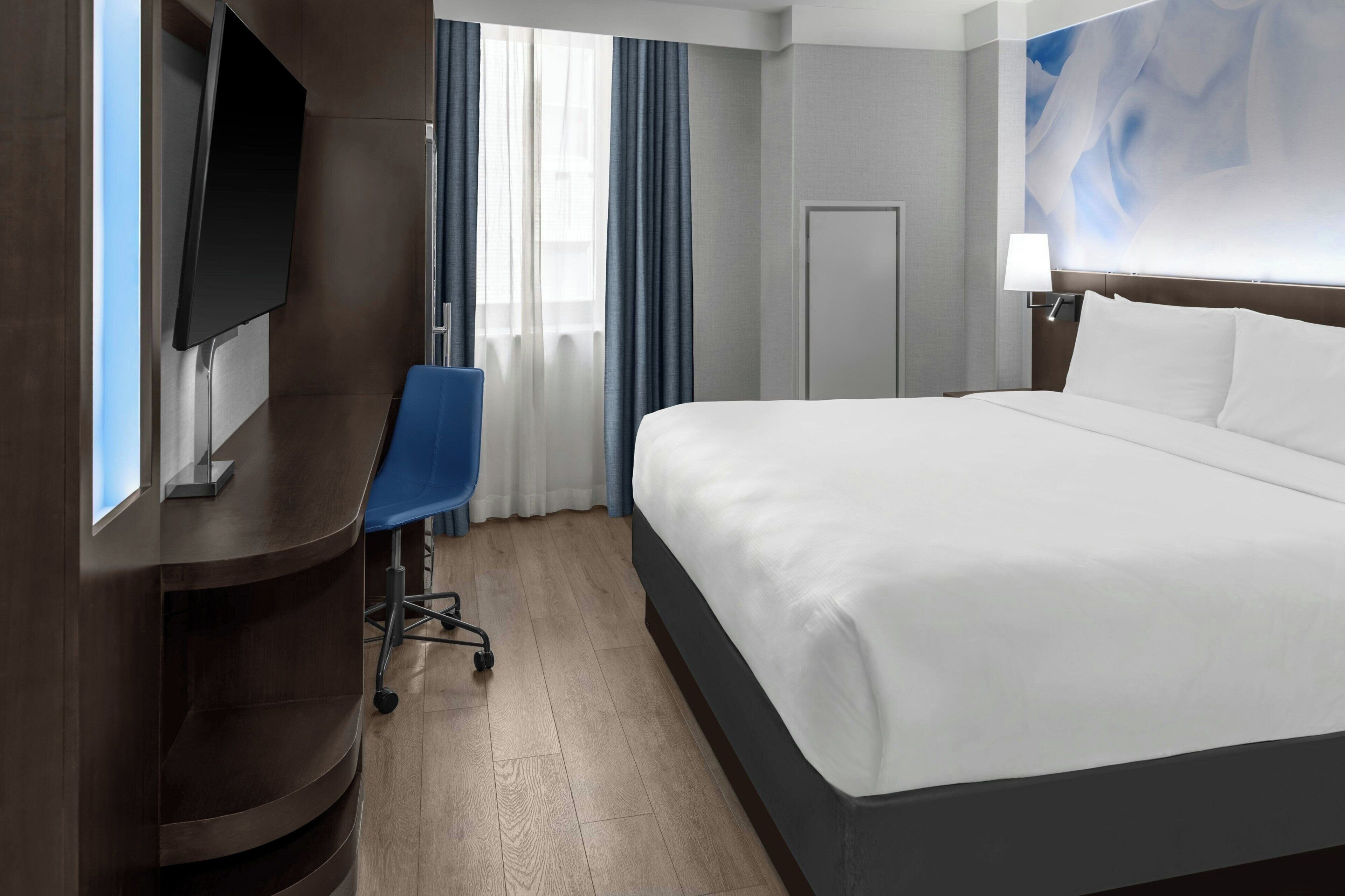 A hotel room with a bed, a desk, a chair, a television, a door, and a window with curtains.