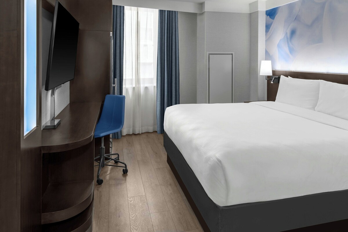 Une chambre d’hôtel avec un lit, un bureau, une chaise, une télévision, une porte et une fenêtre avec des rideaux.