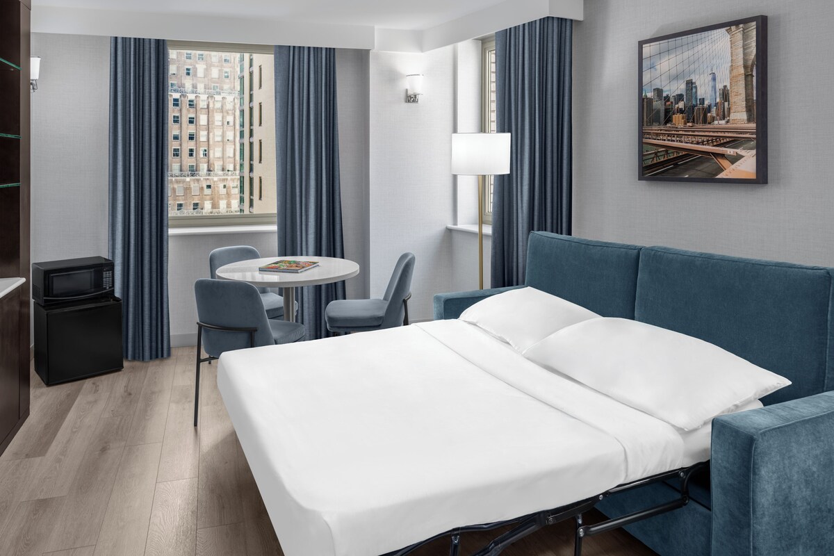 A hotel room with a bed, a desk, a chair, a small table, a lamp, and a cityscape painting.