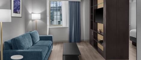 A modern living room with a blue sofa, a small round table, a dark coffee table, a floor lamp, a window with curtains, and a built-in bookshelf.
