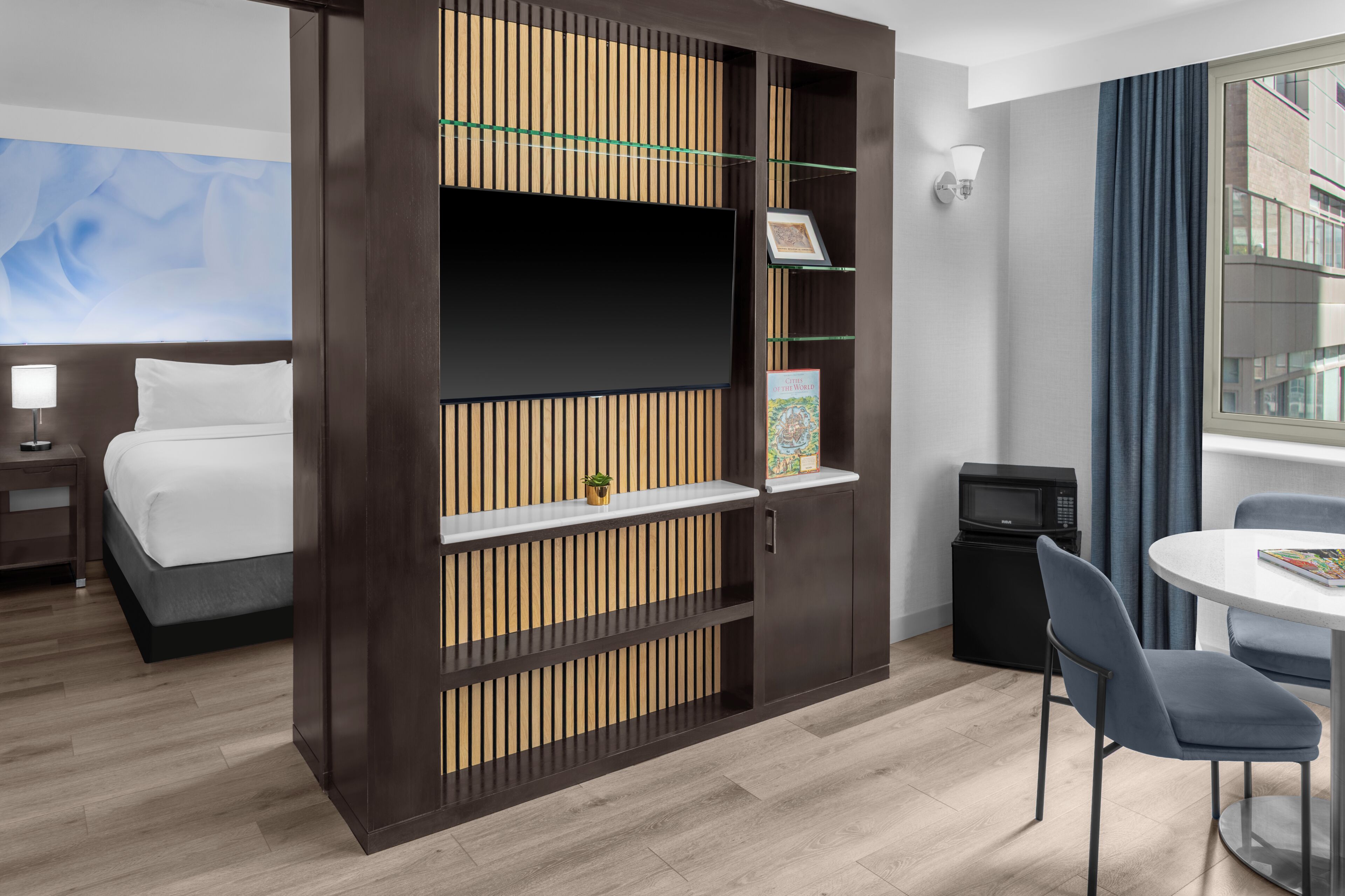 Habitación de hotel con cama, televisor, una mesita con sillas, microondas y una estantería con un marco de fotos.